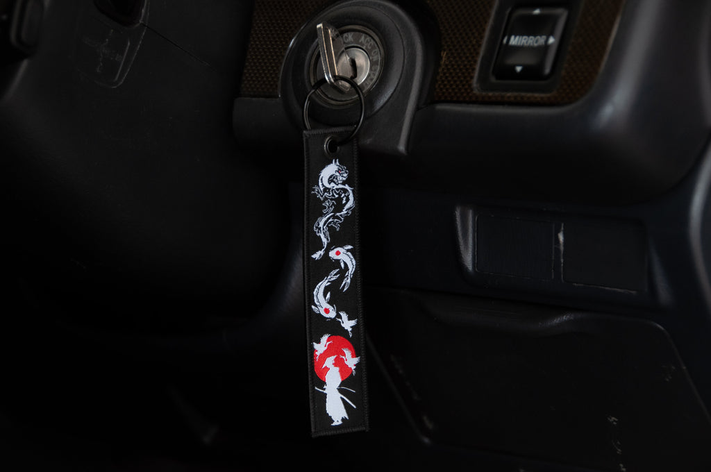 A close-up of a car ignition with a key inserted. Attached to the key is a black jet tag from Incompletegl's The Samurai & His Vision collection, featuring a design with a white dragon and a red sun, incorporating Japanese symbols. The upper part of the dashboard is partially visible in the background.