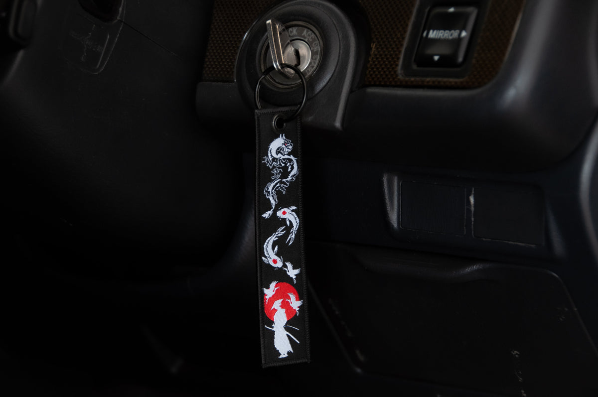 A close-up of a car ignition with a key inserted. Attached to the key is a black jet tag from Incompletegl's The Samurai & His Vision collection, featuring a design with a white dragon and a red sun, incorporating Japanese symbols. The upper part of the dashboard is partially visible in the background.