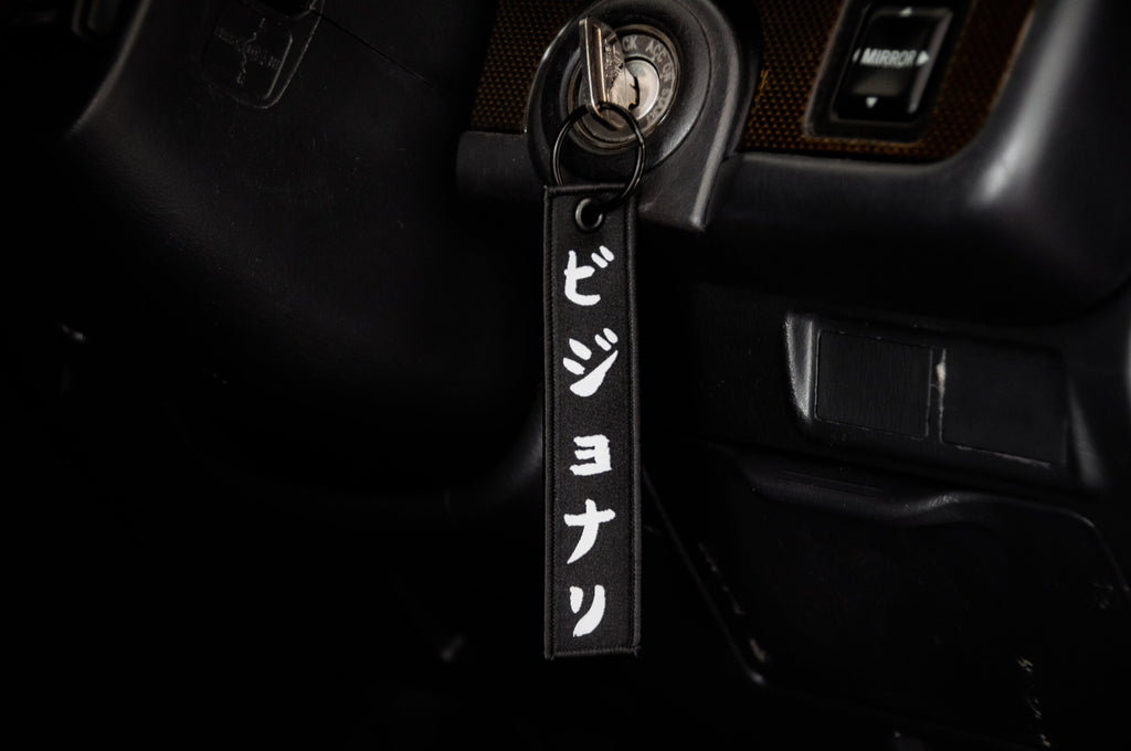 A close-up of a car’s ignition with a key inserted and The Samurai & His Vision | Jet Tag by Incompletegl attached. The black keychain features white Japanese characters representing Japan's most iconic symbols. The car's dashboard and steering wheel are partially visible in the background.