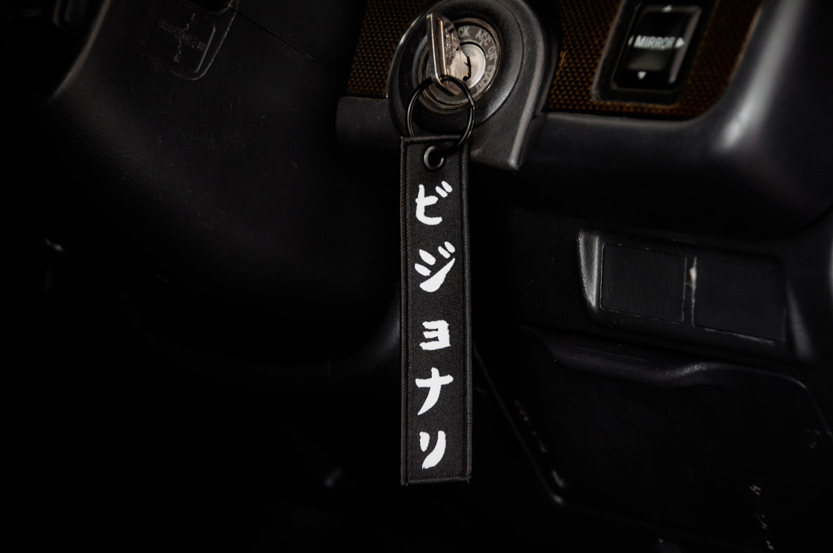A close-up of a car’s ignition with a key inserted and The Samurai & His Vision | Jet Tag by Incompletegl attached. The black keychain features white Japanese characters representing Japan's most iconic symbols. The car's dashboard and steering wheel are partially visible in the background.