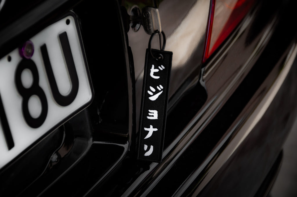 Close-up of a car's rear, featuring a license plate reading "18U" and an Incompletegl "The Samurai & His Vision | Jet Tag" keychain with white vertical Japanese text hanging from the trunk—a nod to Japan's most iconic symbols.