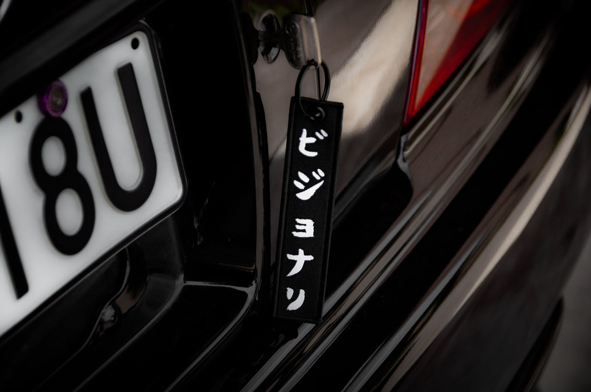 Close-up of a car's rear, featuring a license plate reading "18U" and an Incompletegl "The Samurai & His Vision | Jet Tag" keychain with white vertical Japanese text hanging from the trunk—a nod to Japan's most iconic symbols.