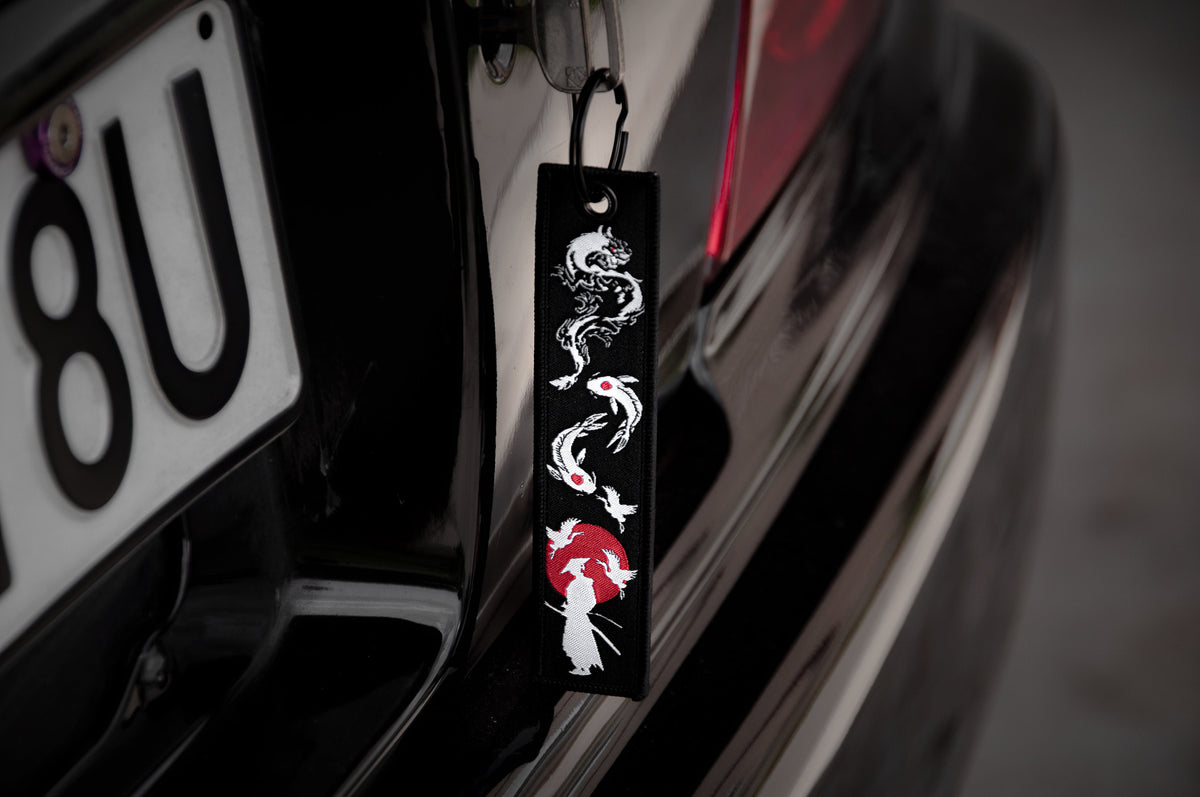 A close-up of a car with "The Samurai & His Vision" Jet Tag by Incompletegl hanging near the license plate. The black fabric strip is embroidered with a white and red dragon and a figure holding an umbrella, echoing Japan's most iconic symbols.