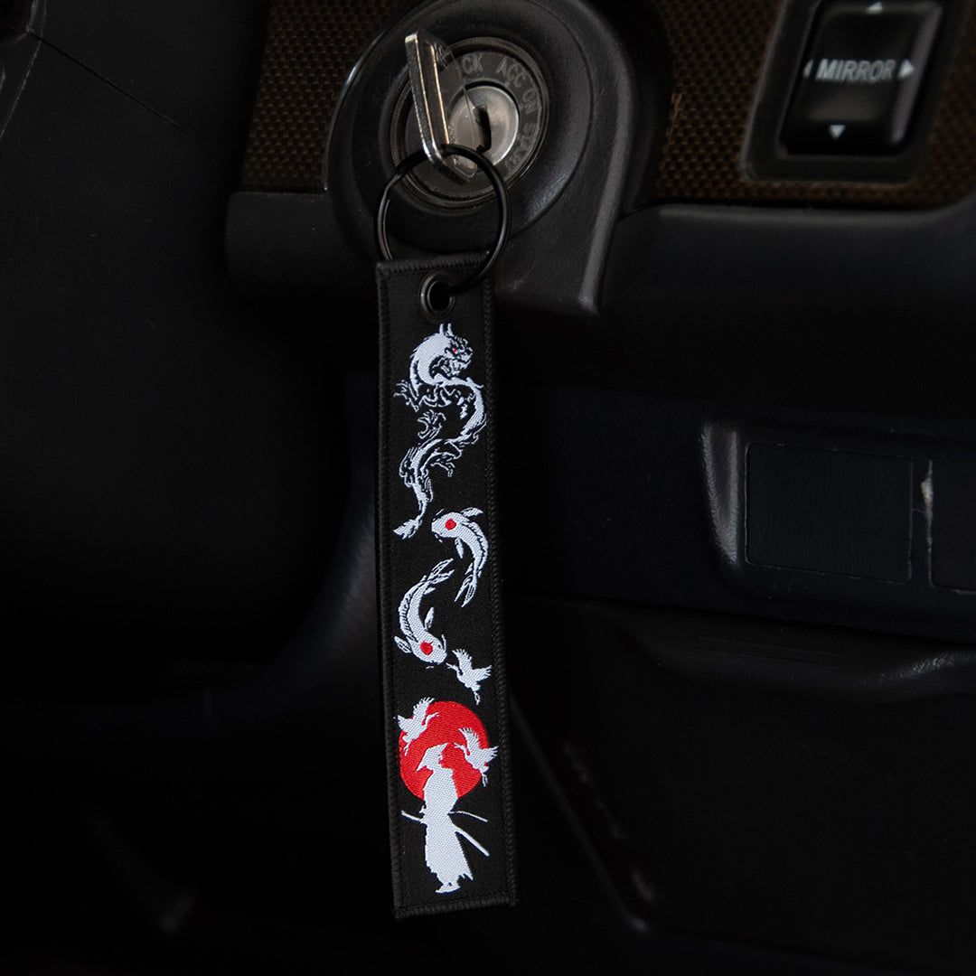 A car key is inserted into the ignition, attached to a rectangular black jet tag named "The Samurai & His Vision" from Incompletegl, featuring a white and red design of koi fish and a red sun—some of Japan's most iconic symbols. The key is in the "lock" position on the vehicle's ignition. The dashboard is also partially visible.